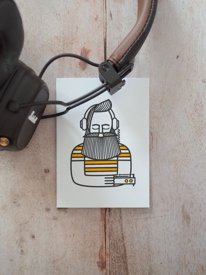 a modern illustration of a man, eyes closed listening to music through headphones is printed onto a small greeting card which sits next to some headphones.