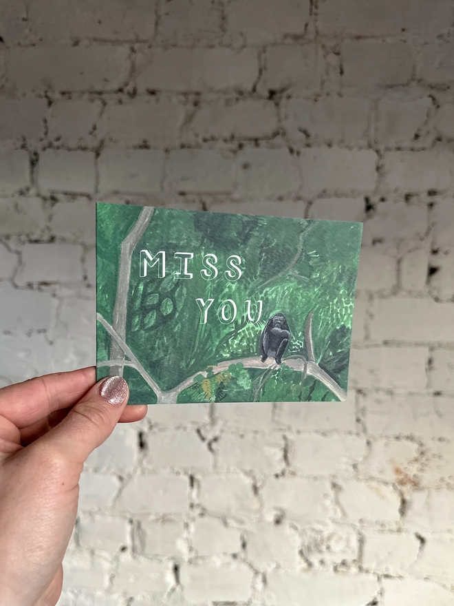 a greetings card featuring an illustration of a chimp sitting in a jungle landscape with the text “miss you”