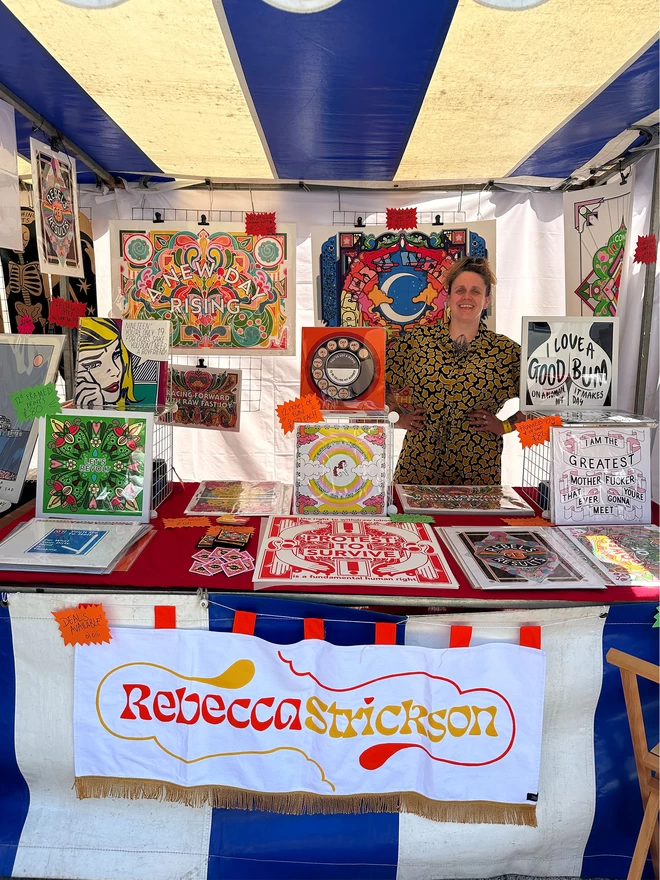 The artist stands at a market stall displaying an array of her artworks, and a banner with her Rebecca Strickson logo