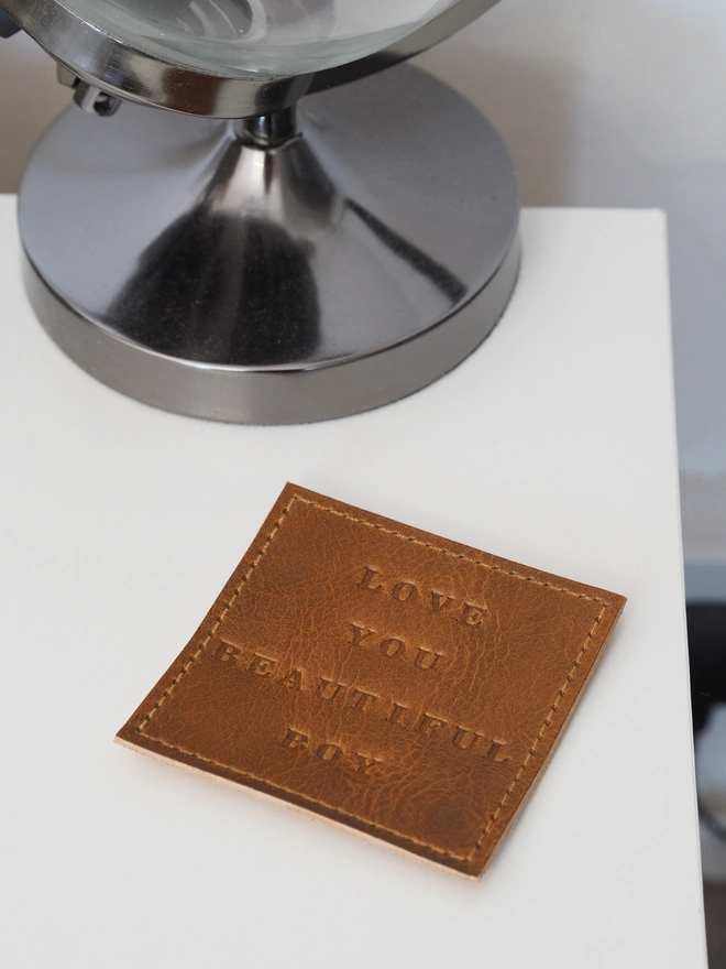 Personalised leather coaster. Hand dyed tan brown square leather coaster, sitting on a bedside table. Large print hand stamped letters spell out a short but sweet message.