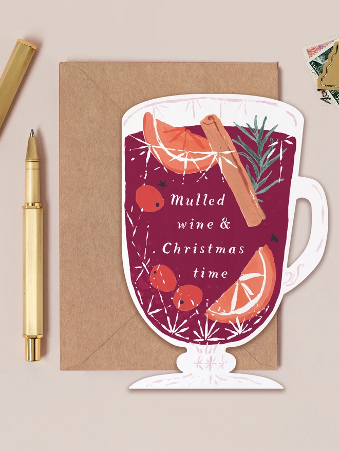 Mulled Wine Christmas card shaped like a glass mug of mulled wine with orange slices and cinnamon, lettered 'Mulled wine & Christmas time', on a kraft envelope beside a gold pen