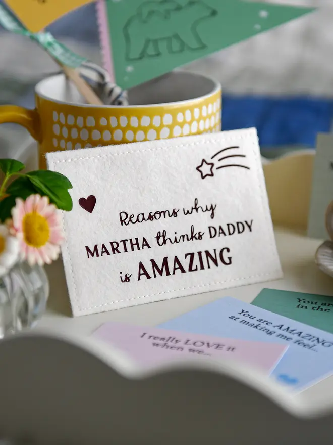 A close-up of a handmade felt envelope with the words "Reasons why MARTHA thinks DADDY is AMAZING" displayed on a white breakfast tray. Around it are pastel-coloured prompt cards with handwritten messages, a small vase of daisies, a yellow mug with crafting items, and a miniature candle on a ceramic plate, creating a cosy Father's Day setting.