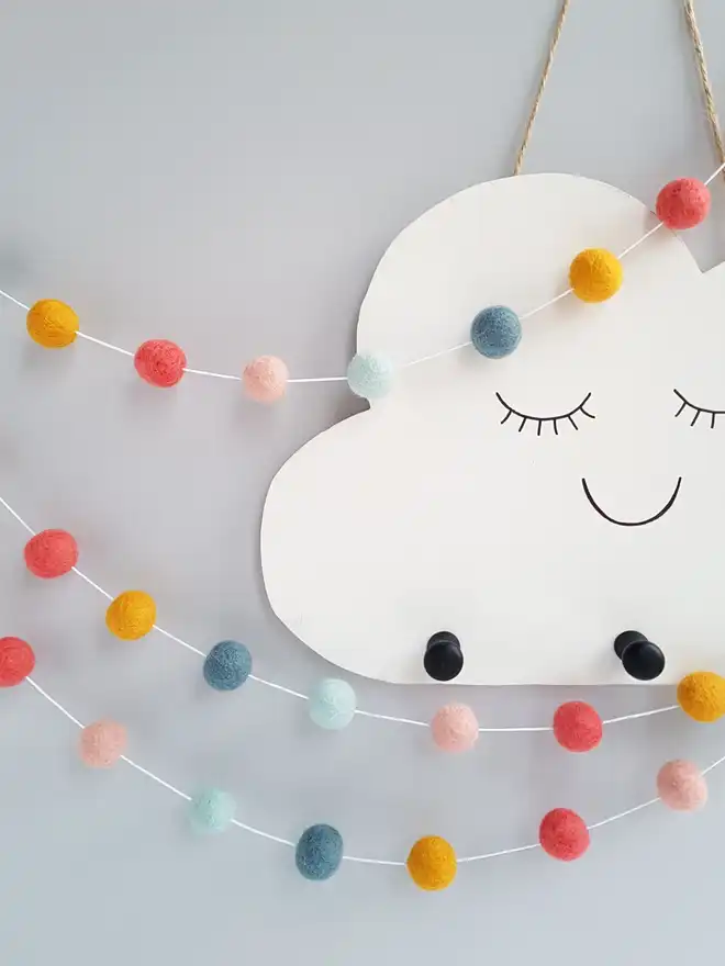 A pastel rainbow felt ball garland hanging in front of a decorative white cloud.