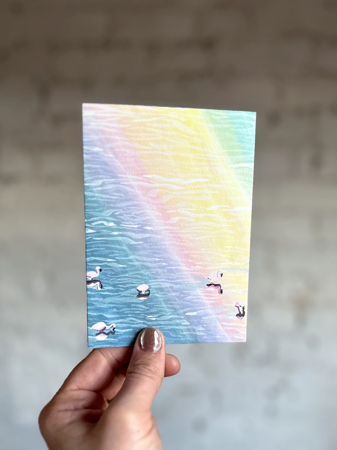 a greetings card featuring an illustration of flamingos in water with a rainbow reflection