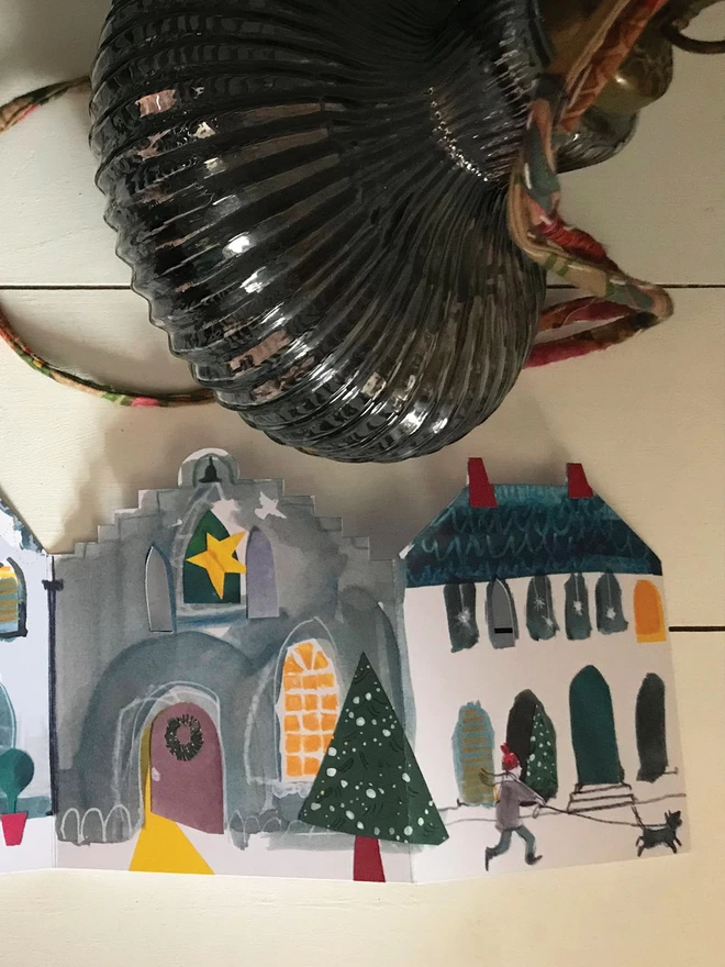 A detail of a Festive Street illustrated concertina greetings card, lying against a painted board background, alongside a vintage glass bauble.