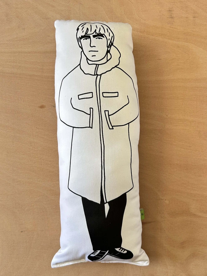 Black and white illustrated screen printed cushion of Oasis band member Liam Gallagher. The cushion sits on a light wooden surface.