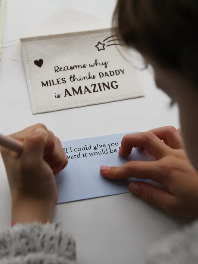 A child’s hands are shown writing on a light blue prompt card that says “You are AMAZING at making me feel…” using a green pencil. Other completed cards with handwritten messages are visible nearby on a white desk, including one that says “The best adventure we’ve had together was…” This scene captures a child filling out a sentimental Father’s Day gift.