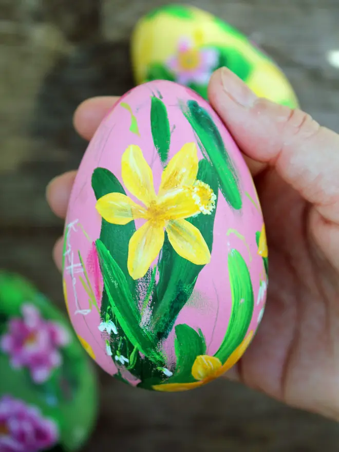 Hand painted floral wooden egg. pale pink background adorned in yellow daffodils and white snowdrop flowers. Held in a woman's hand two other eggs in the background out of focus.