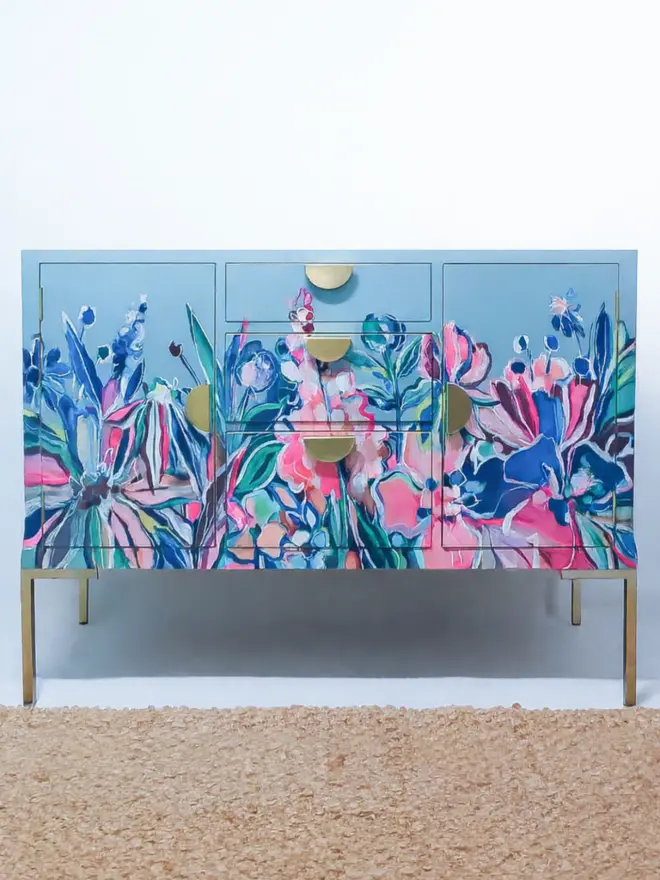 Beautiful Vintage remodelled sideboard with brand new brass hardware inclusions half-moon handles and legs. The sideboard has a central bank of 3 drawers with a cupboard space either side and is painted in dusky blue eggshell paint. The front of the sideboard features a hand-painted frieze by Chloe Kempster with peonies, daisies and abstract floral shapes in neon pink, green, navy blue, pale pink and an assortment of other colours. 