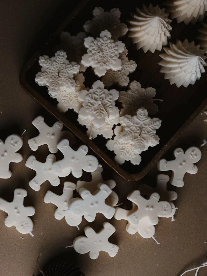 White Gingerbread Candles (Set Of 2). A top-down view of cream gingerbread man candles scattered on kraft paper beside a wooden tray of embossed snowflake candles and swirled tree candles. The playful festive shapes make charming stocking fillers, table favours or Secret Santa gifts.