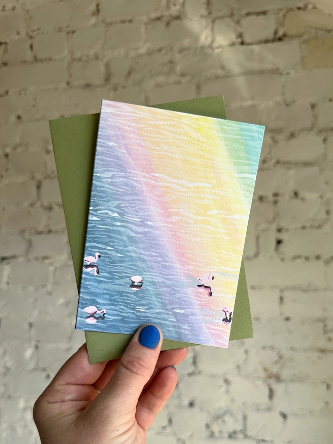 a greetings card featuring an illustration of flamingos in water with a rainbow reflection with a green envelope 