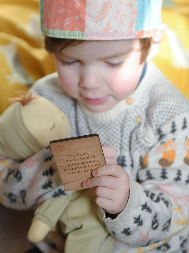 A young toddler wearing a patterned jumper and a fabric crown holds a wooden story card in one hand and a soft doll in the other. 