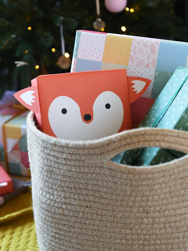 A gift wrapped as an orange fox is tucked into woven basket beside other wrapped gifts in front of a Christmas Tree.