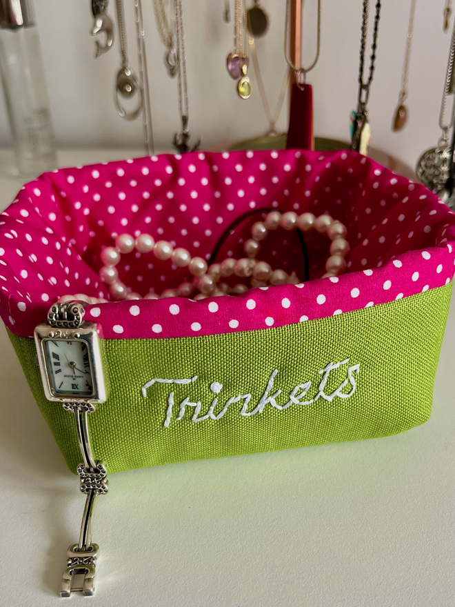 Green fabric basket with pink polka dot lining, embroidered with ‘Trinkets’ & holding jewellery.