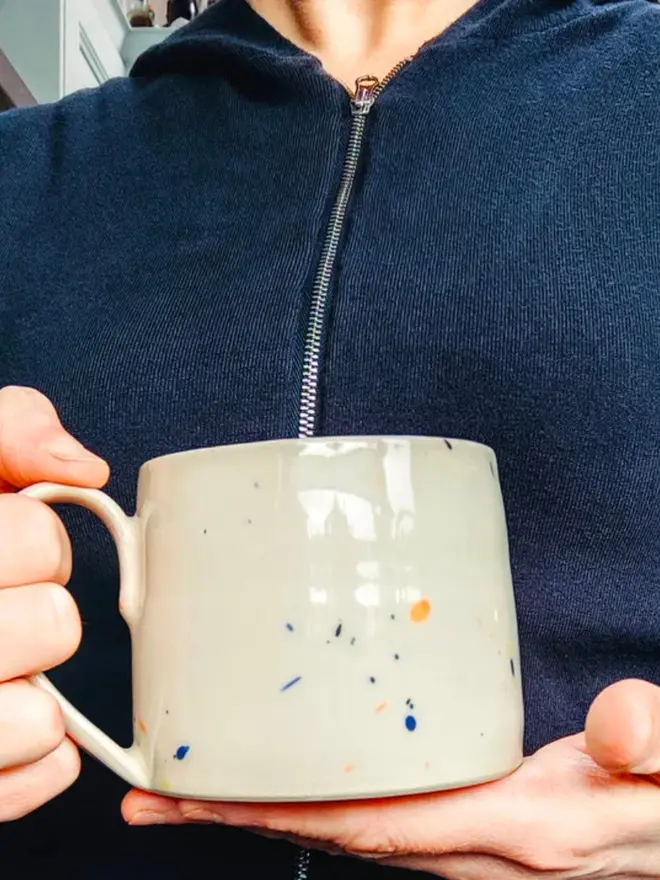 Large creamy white terrazzo cups with black, blue, and yellow splatters and glossy glaze being held by a person wearing a black top.