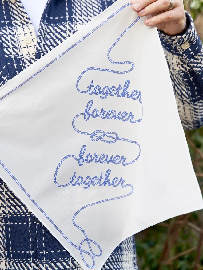 Together Forever white cotton handkerchief held up in a garden setting by a man with a white and blue checkered jacket