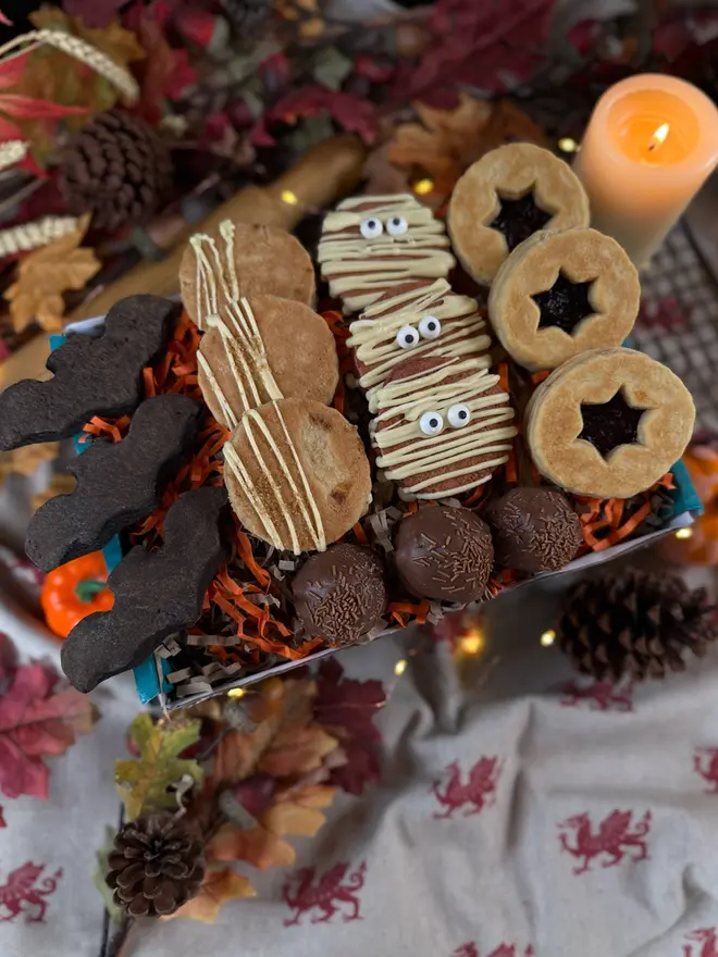 A Halloween-themed welshcake box filled with assorted treats, including red velvet mummy welshcakes, star-shaped blackberry sandwich welshcake, chocolate balls, and bat-shaped black velvet welshcake, and apple crumble welshcake arranged in orange shredded paper inside a blue box on a table with a candle and autumn leaves in the background.