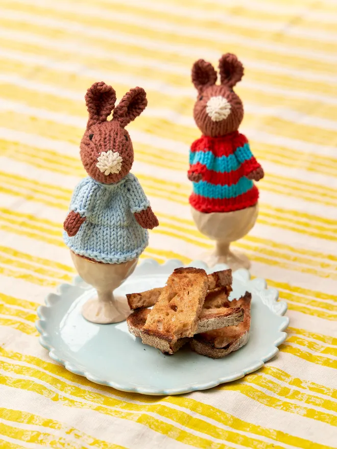 Egg Cosy Rabbits Easter Table Decoration, a pair of colourful Easter cosies sitting on a striped tablecloth surface. 