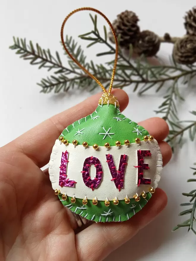 A Hetty and Dave green hand stitched leatherette retro inspired bauble with pink metallic lettering spelling out LOVE on a white band, adorned with tiny gold beads
