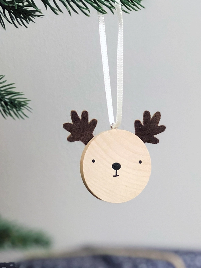 A small wooden and felt reindeer decoration hangs on a Christmas tree branch.