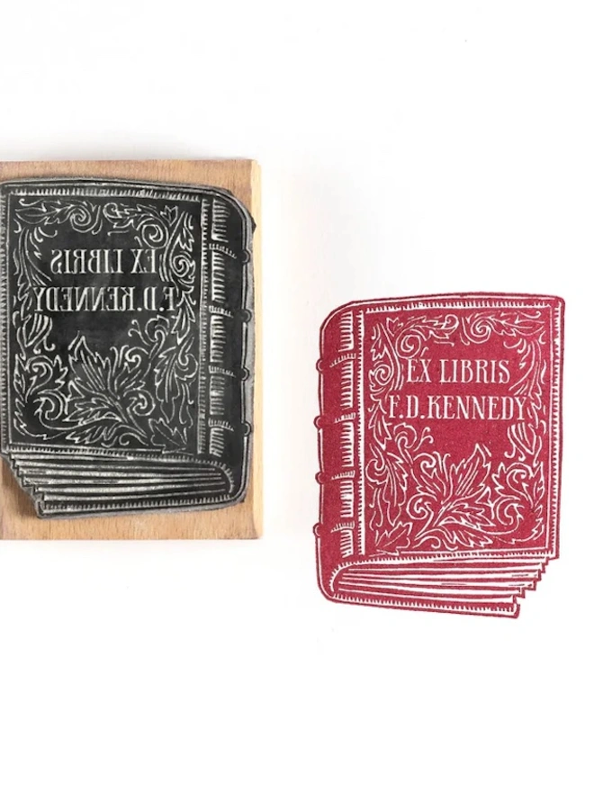 Wooden rubber stamp with an illustrated antique book design, shown next to a red ink stamped impression reading ‘Ex Libris F. D. Kennedy’.