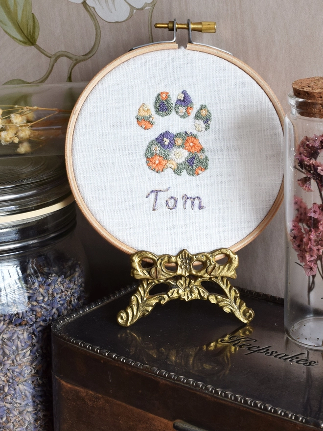 An embroidered Cats Paw of Golden Yellows and Bright Orange Blossoms with Green French Knot grass background.  Displayed in a hoop frame on a Gold tone stand, on top of a tarnished silver keepsake tin with floral specimen glasses.