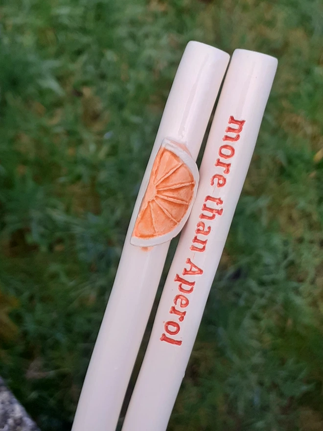 Handmade ceramic drinking straw set on a grassy background, featuring a raised orange slice decoration and a second straw with red text reading “more than Aperol.”