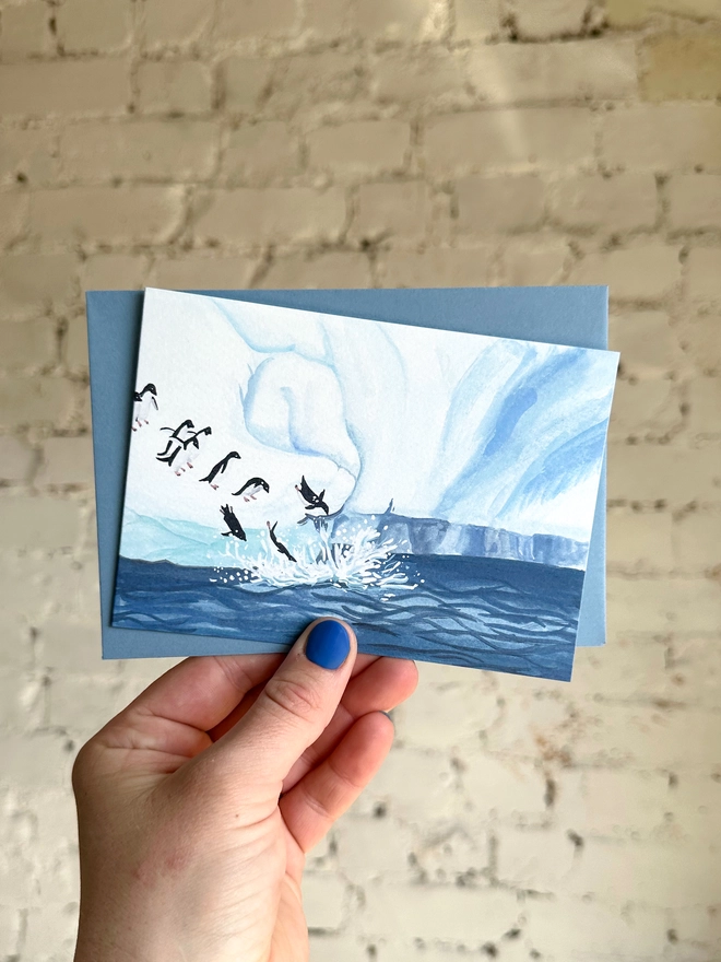  a greetings card featuring an illustration of an icy landscape with penguins diving into the sea with a blue envelope
