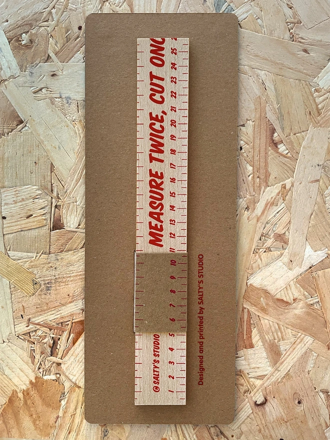 A wooden ruler in a brown cardboard display pack, is vertical in the frame, sat on a chip board background. Measure Twice Cut Once is printed onto the ruler as a joke, part of the phrase is missing. Cut off.