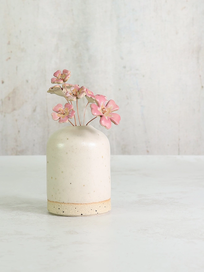white ceramic handmade vase sits on a white table top with a posy of pink ceramic primroses inside. The background is soft grey tones 