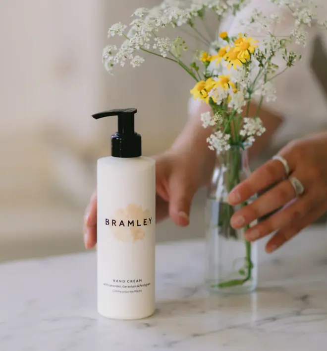 person holding a bottle of white hand cream with a black pump lid, and a glass vase of yellow and white flowers