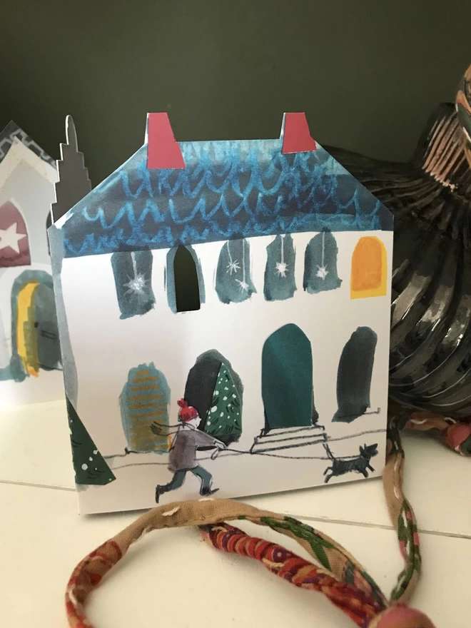 Festive Street illustrated concertina card showing a festive house and dog walker, sits on a shelf alonside a vintage glass bauble.