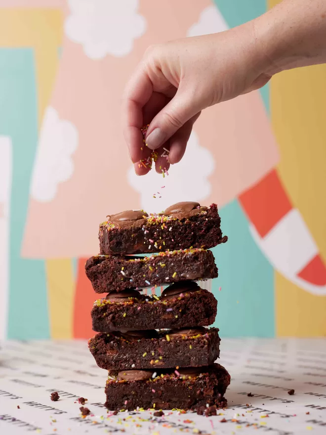 Funfetti Brownie stack of five slices with sprinkles and milk chocolate buttons and a hand sprinkling Funfetti on top against a colourful backdrop