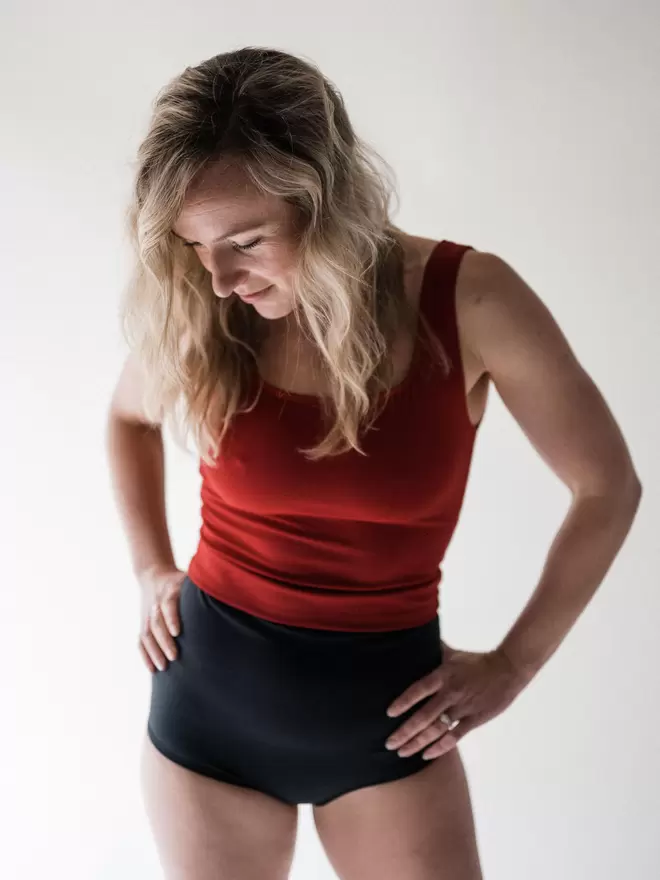 Blonde woman looking downwards in studio with hands on hips wearing Davy J Sustainable Waterwear red crop swim top overlapping black high waist bikini briefs