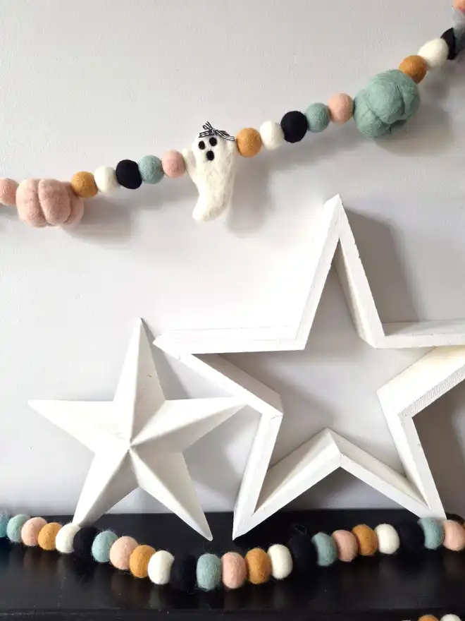 Two stars on a black mantelpiece, with a felt ball garland laid on top. Plus a ghost and pumpkin garland hanging above in soft, neutral shades.