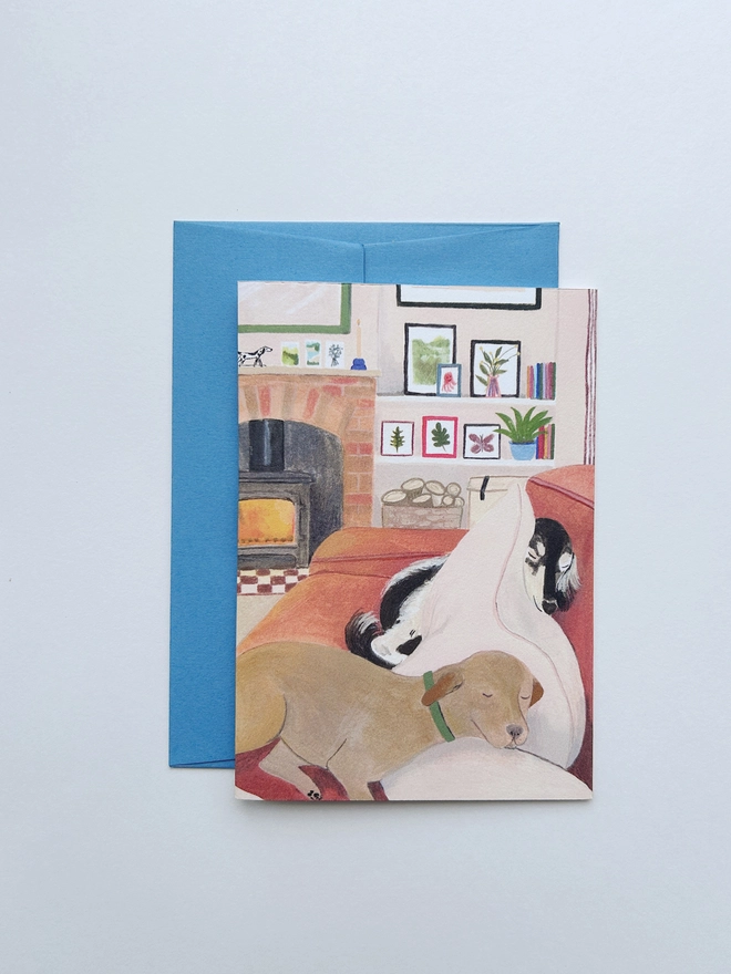 A greeting card showing two dogs cuddled up on the sofa together, next to a roaring fire. The dogs are a black and white saluki and a fox red labrador