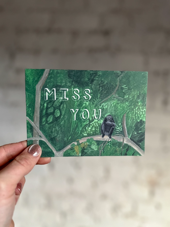 a greetings card featuring an illustration of a chimp sitting in a jungle landscape with the text “miss you”