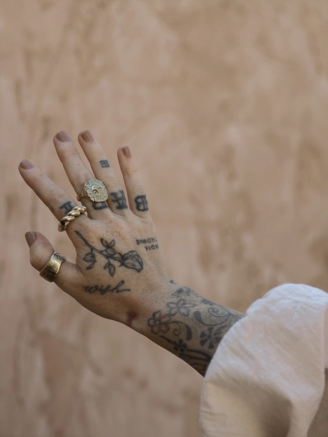 The image of a tattooed female hand wearing a hand carved gold toned brass sun face ring, a twisted gold toned brass ring with a blue sapphire and a thick cigar style gold toned brass ring with star set crystals in front of a rustic terracotta coloured plaster wall
