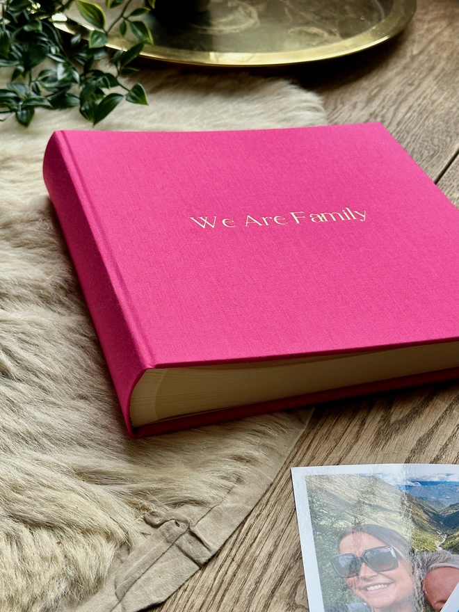 A hot pink photo album printed with 'We Are Family' in gold lettering sits on a wooden surface, next to a pile of photos.