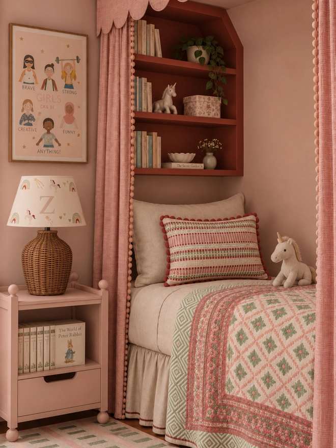 Studio Zef personalised unicorn lampshade with pastel rainbows, unicorns and the letter Z, styled on a wicker table lamp beside a cosy pink canopy bed with patterned bedding and built-in bookshelves in a girls' bedroom.
