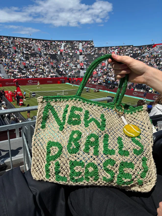 'New Balls Please!' Bag in front of a grass tennis court.