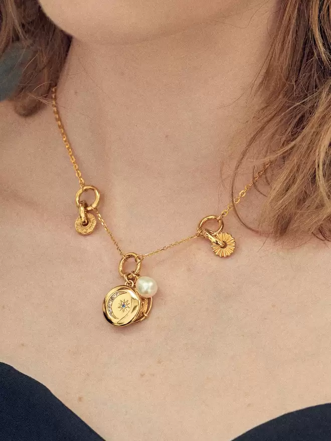 Woman wearing a gold necklace styled with a gold locket, a pearl amulet and two gold disc charms