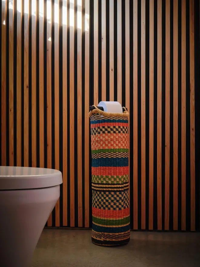 Ghanaian Hand Woven Tubular Toilet Roll Holder, a toilet roll holder sitting in a bathroom. It sits in front of a wooden backdrop. 