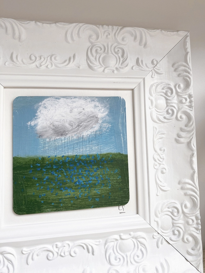 wooden shelf with painting of blue sky, a cloud and a field of blue forget-me-nots in chunky white ornate frame