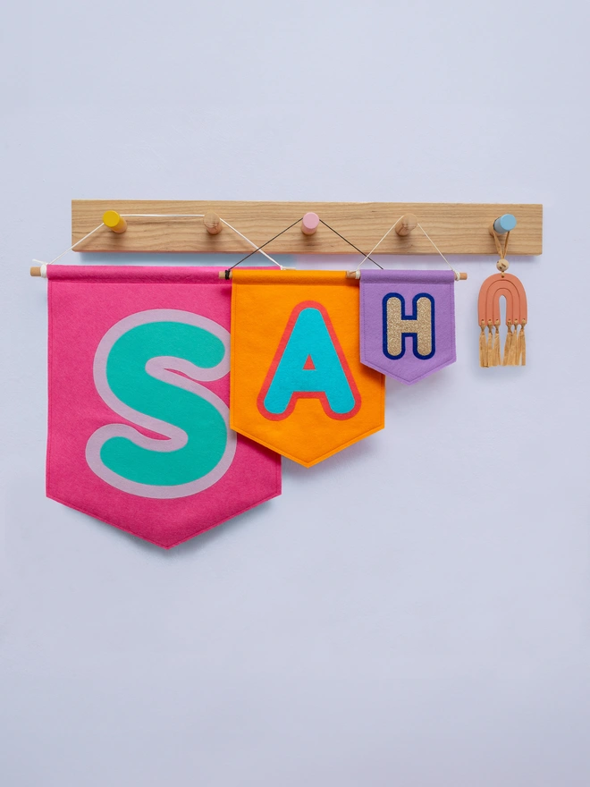 3 brightly coloured banners hanging from a wooden peg rail. The banners are 3 different sizes and have initials on the. Each initial is surrounded by a contrasting coloured bubble
