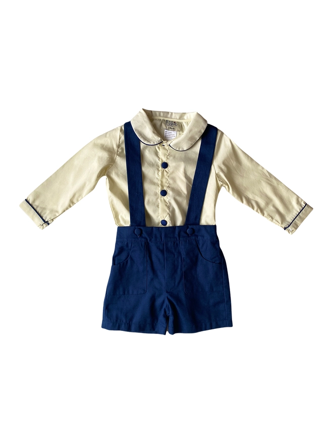 Cutout of cream shirt with navy piping and buttons and navy shorts with braces