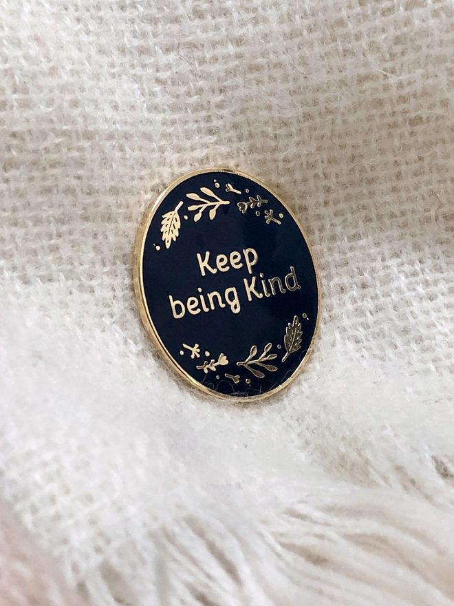 A navy blue and gold enamel pin  with the words Keep Being Kind with a floral border is pinned onto a white scarf.