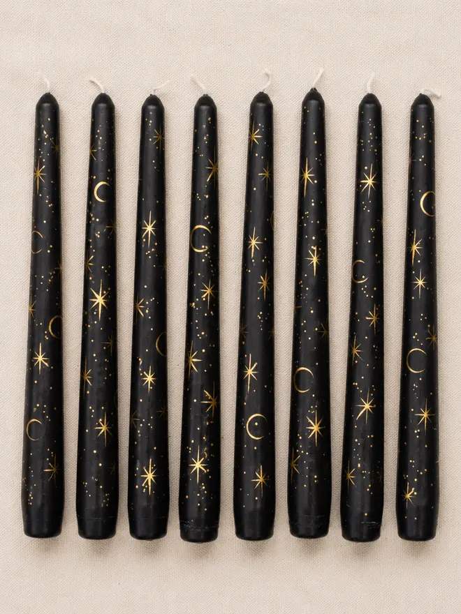 Eight hand-painted black taper candles decorated with gold moons and stars, arranged in a neat row on natural linen fabric.
