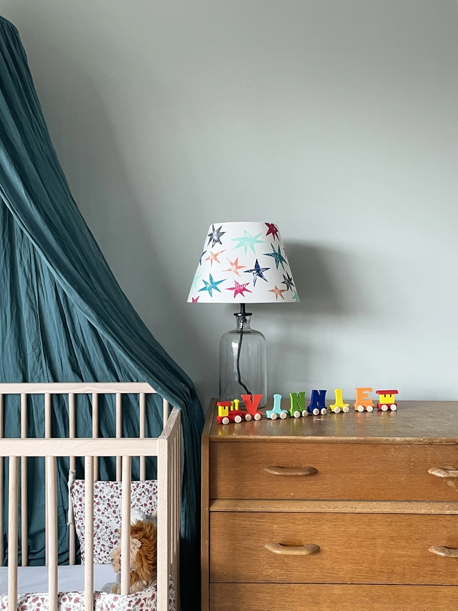 A colourful lampshade with multicoloured star prints, styled on a glass base place on a wooden chest of drawers next to a cot. This empire shade for table lamps is a cheerful lampshade with stars, perfect for kids bedroom decor.