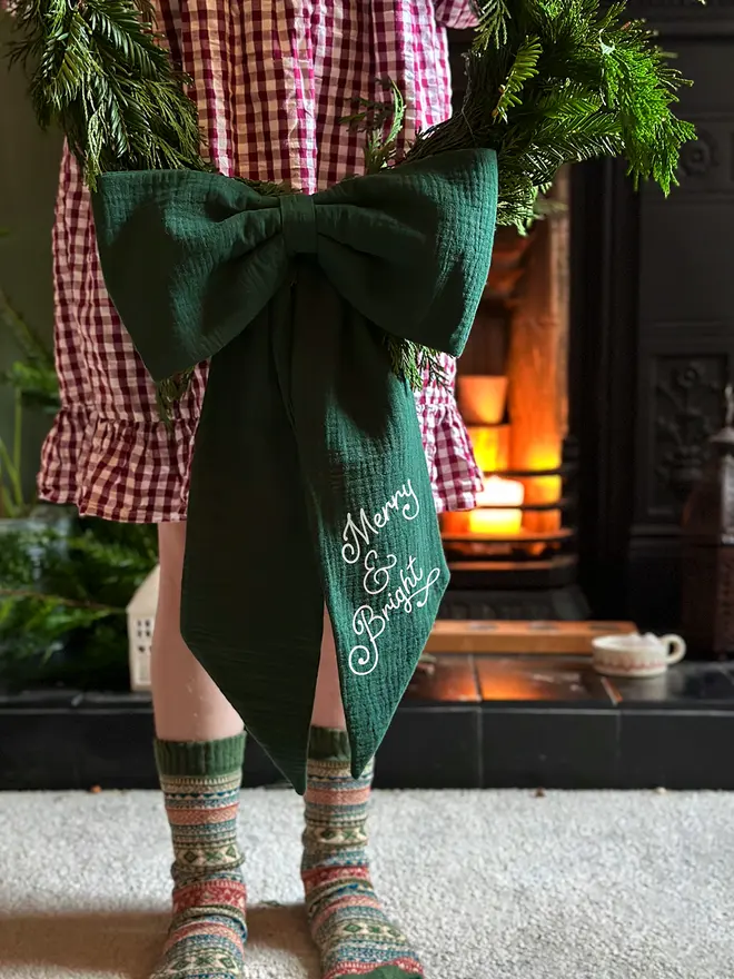 A handmade fabric 'Merry and Bright' Christmas bow decoration made from forest green cotton is being held on a Christmas wreath by a child wearing a red gingham dress.